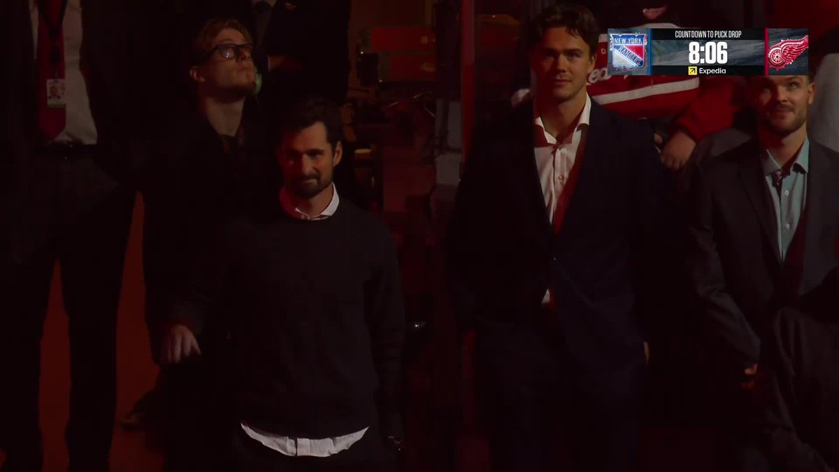 Three men in suits and sweaters standing together at what appears to be a hockey arena, with a broadcast graphic in the corner showing 'COUNTDOWN TO PUCK DROP 8:06' between the Rangers and Red Wings.