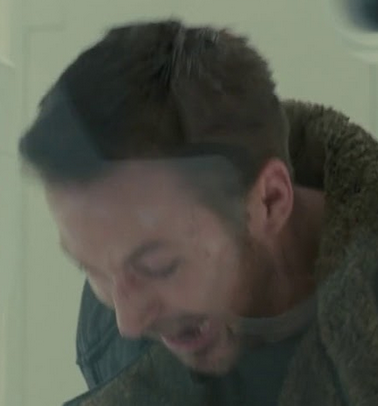 A grainy close-up of a bearded man in a heavy coat, appearing injured and looking downward in a dimly lit scene.
