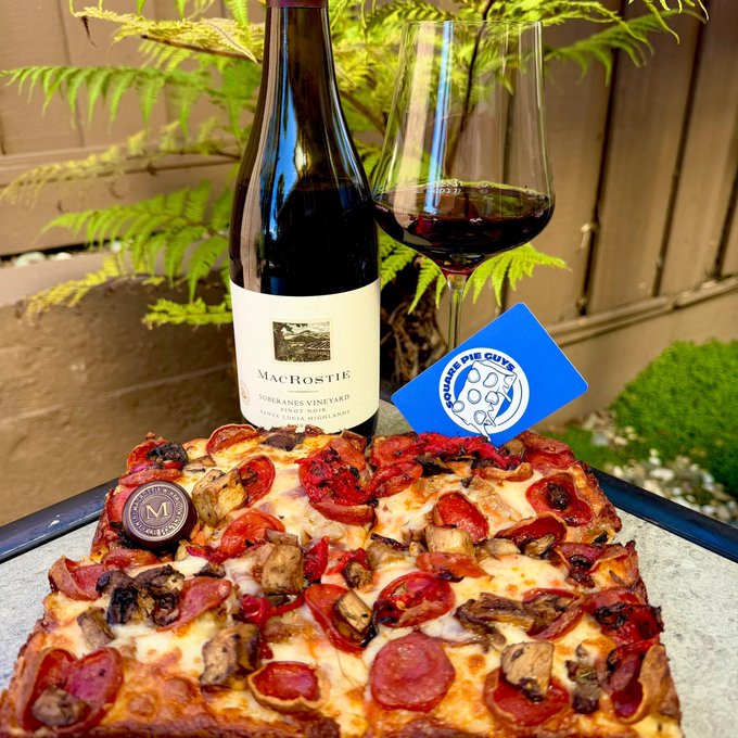 A bottle of MacRostie Sonoma County Pinot Noir red wine paired with a glass and a pizza topped with tomatoes, meat, and cheese, accompanied by a blue 'Square Pie Guys' napkin.