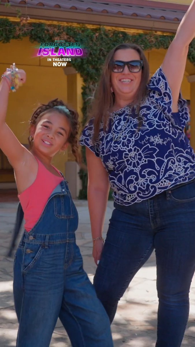 A woman and young girl pose cheerfully with raised arms in front of a yellow building, both wearing denim overalls, with a "FORGOTTEN ISLAND IN THEATERS NOW" promotional graphic in the corner—illustrating the post's theme of families making memories at the movie.