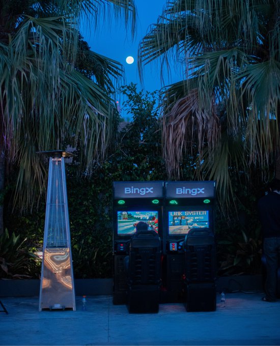 An outdoor evening scene featuring two arcade-style gaming stations branded 'BingX' displaying 'LINK SYSTEM' alongside a pyramid patio heater, set against a backdrop of palm trees and a visible full moon.