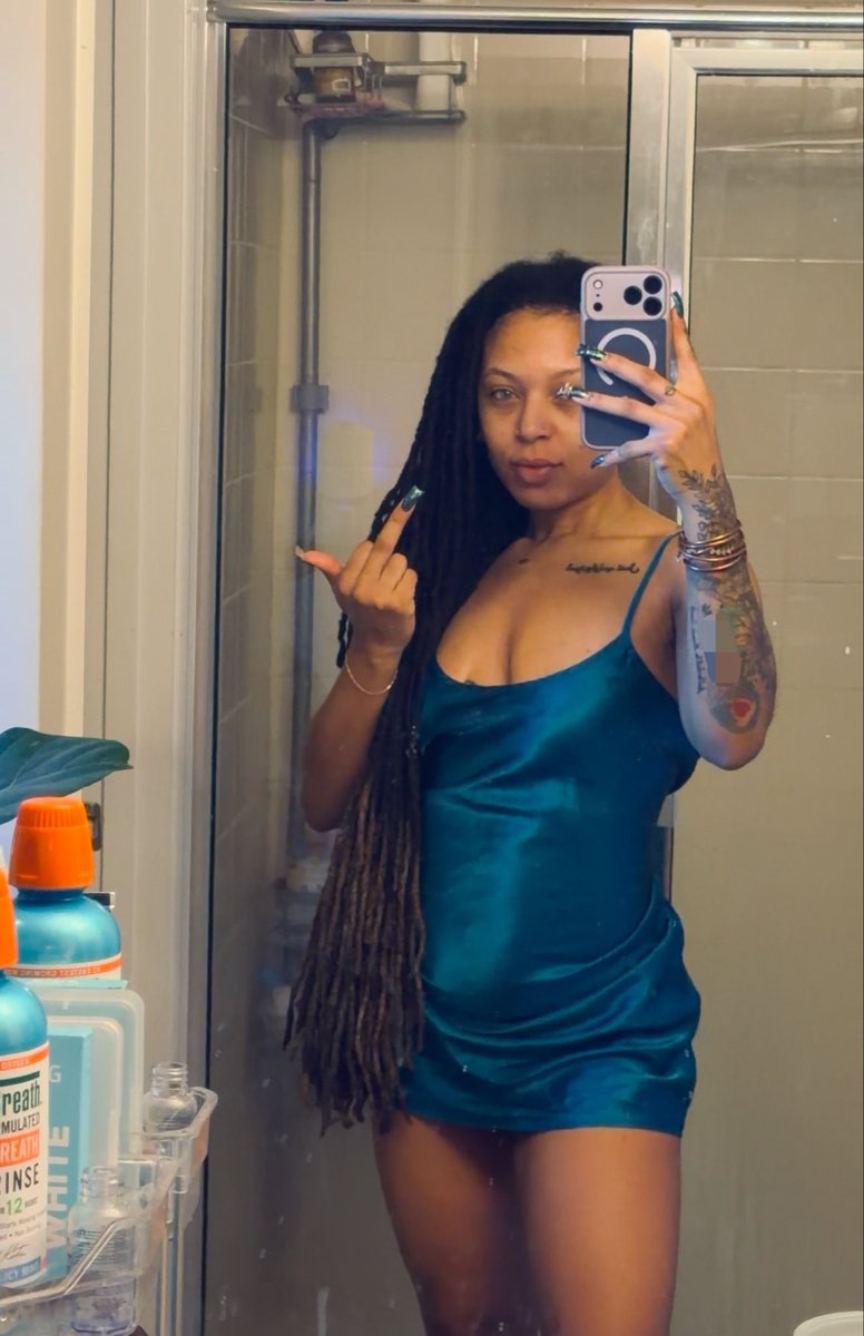 A woman with long dark dreadlocks wearing a teal satin slip dress takes a mirror selfie in a bathroom, holding a phone with a magnetic ring and making a middle finger gesture with her other hand.