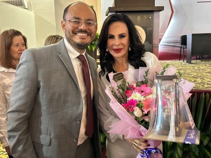 A man in a gray plaid suit and a woman in a light-colored outfit posing together at an event, with the woman holding a pink bouquet and a boxed Loumir award/trophy.