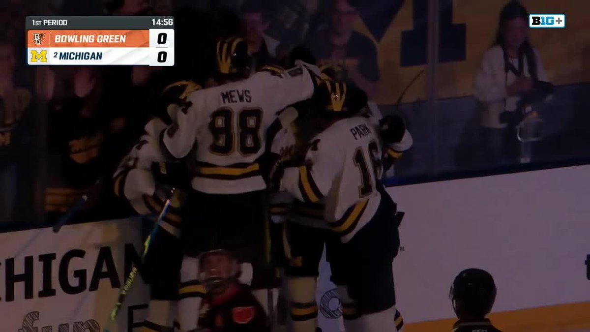 A hockey broadcast showing players in white and yellow jerseys (including #88 MEVS and #19) celebrating together against the boards, with a scoreboard indicating Bowling Green 0 vs. #2 Michigan 0 at 14:56 of the 1st Period on B1G+.