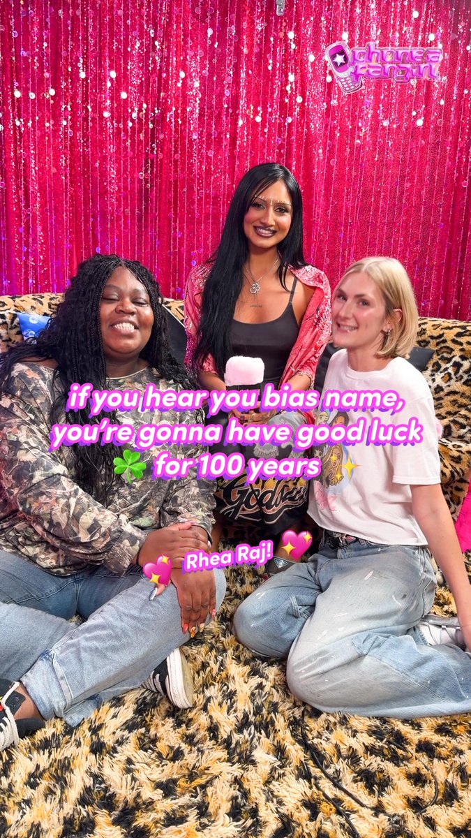 Three women pose together on a leopard-print couch in front of a pink sequined backdrop. Text overlay reads 'if you hear you bias name, you're gonna have good luck 🍀 for 100 years ✨💫', 'Rhea Rai! 💖✨', and includes a 'Phone a Friend' logo in the corner.