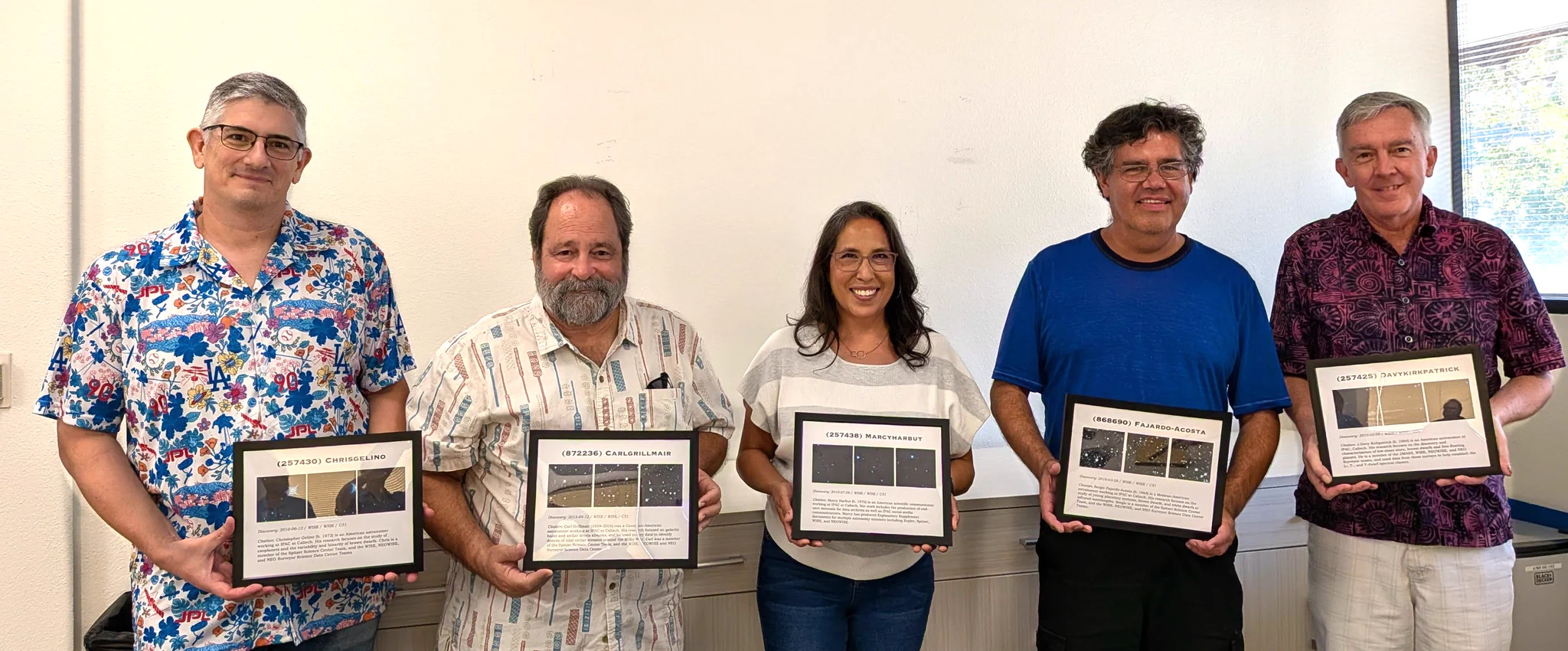 This is a group photo of five people standing against a white wall and looking and smiling at the camera while each holding a plaque of their Minor Planet designation. The individuals are (from viewer's left to right):  Chris Gelino, Roc Cutri (holding Carl Grillmair's plaque), Marcy Harbut, Sergio Fajardo-Acosta, and J. Davy Kirkpatrick. 