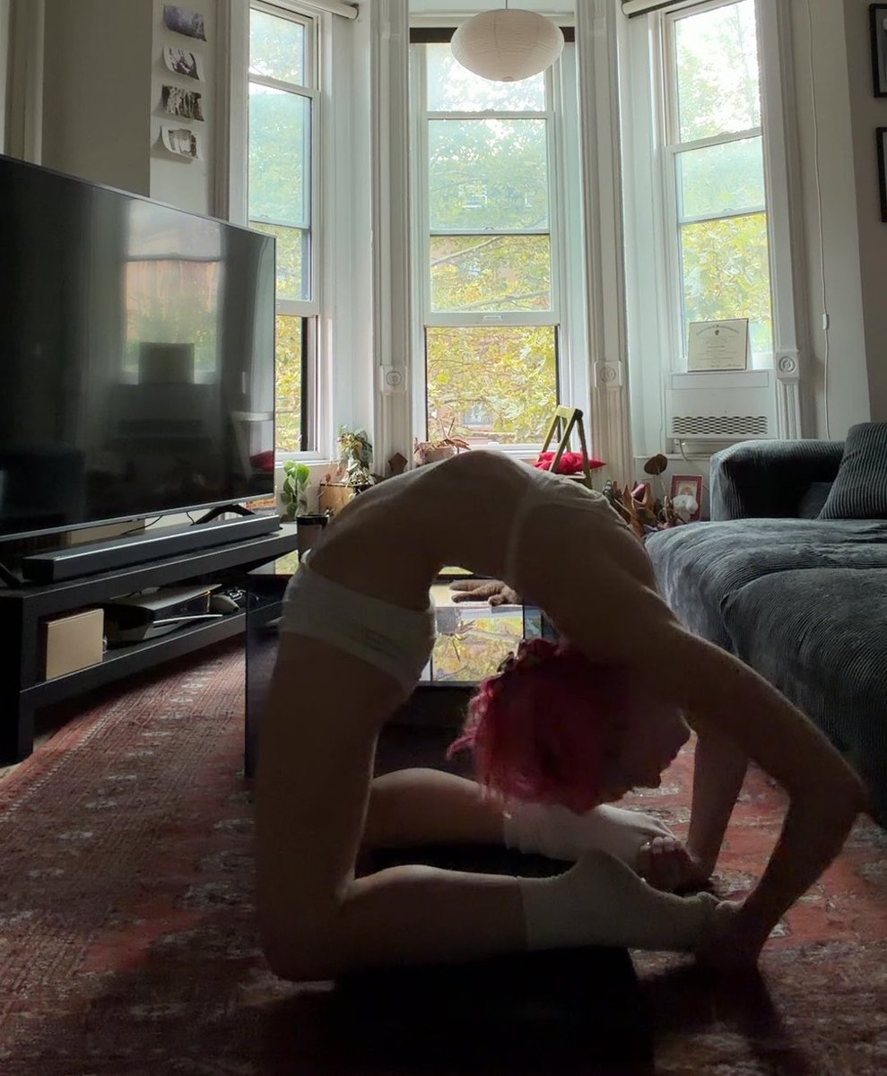 A young person with pink hair performs an impressive backbend contortion in a sunlit living room, wearing white leg warmers and a leotard. The flexible pose—with chest lifted and hands grasping feet behind the head—reflects the caption "Triplefold soon I reckon," suggesting advanced contortion training in progress.