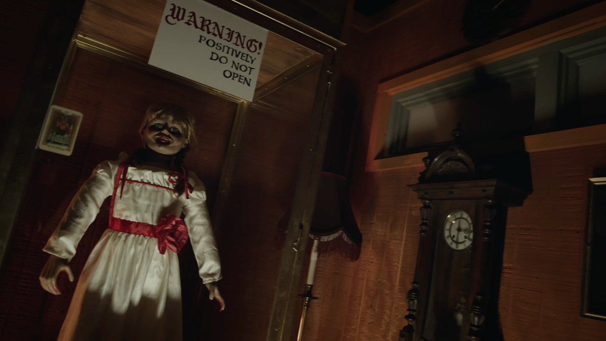 A creepy doll with a cracked face and white dress with red ribbons stands inside a glass case beneath a sign reading "WARNING! POSITIVELY DO NOT OPEN," next to an old grandfather clock, capturing the haunting atmosphere of a haunted house attraction.