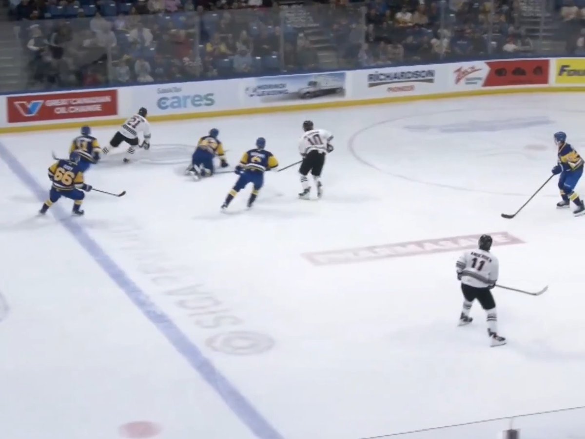 An overhead view of an ice hockey game in progress with players in blue/yellow and white/black jerseys on the ice, with advertisements visible on the boards including 'great canadian oil change' and 'sasktel cares'.