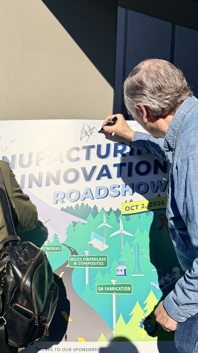 A man in a denim shirt signs a large colorful event poster while another person with a backpack stands beside him. The poster features bold lettering and illustrated scenery, with additional text.