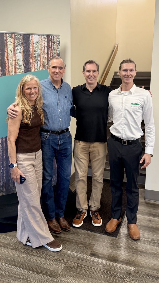 A posed indoor photograph of four smiling adults standing close together with their arms around one another. A forest mural, staircase, and wood-look flooring are visible in the background. There is a small name badge with text on one person's shirt.