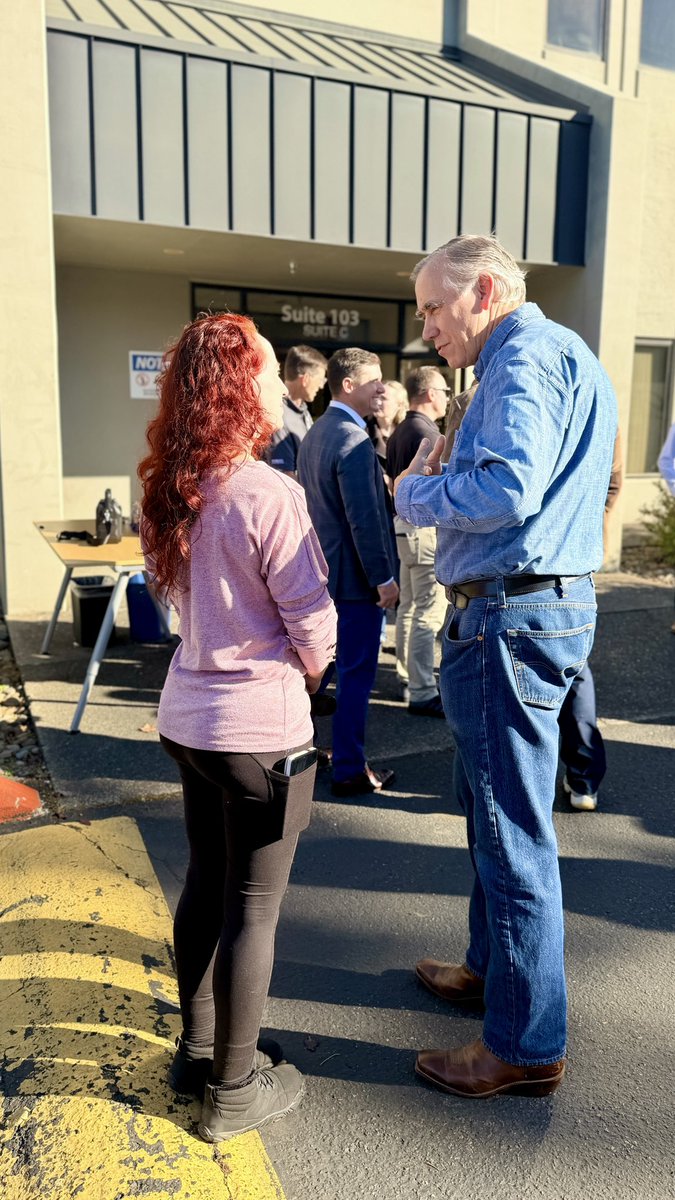 People are gathered outside a commercial building in bright sunlight. In the foreground, a woman and an older man talk while several people stand near the entrance behind them. A folding table and chairs are set up nearby. There is visible text on the building and a sign.