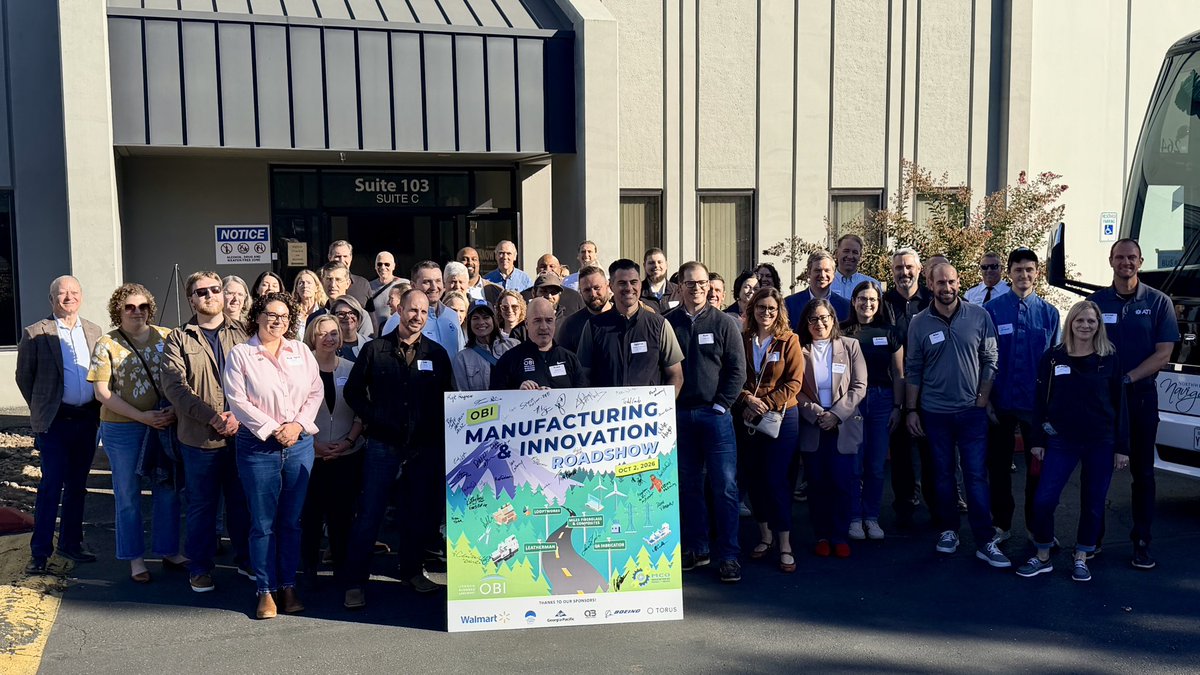 A large group of adults poses outside a commercial building, with a colorful event poster displayed in front of them. A vehicle is partially visible at the right. There is text on the building and poster.