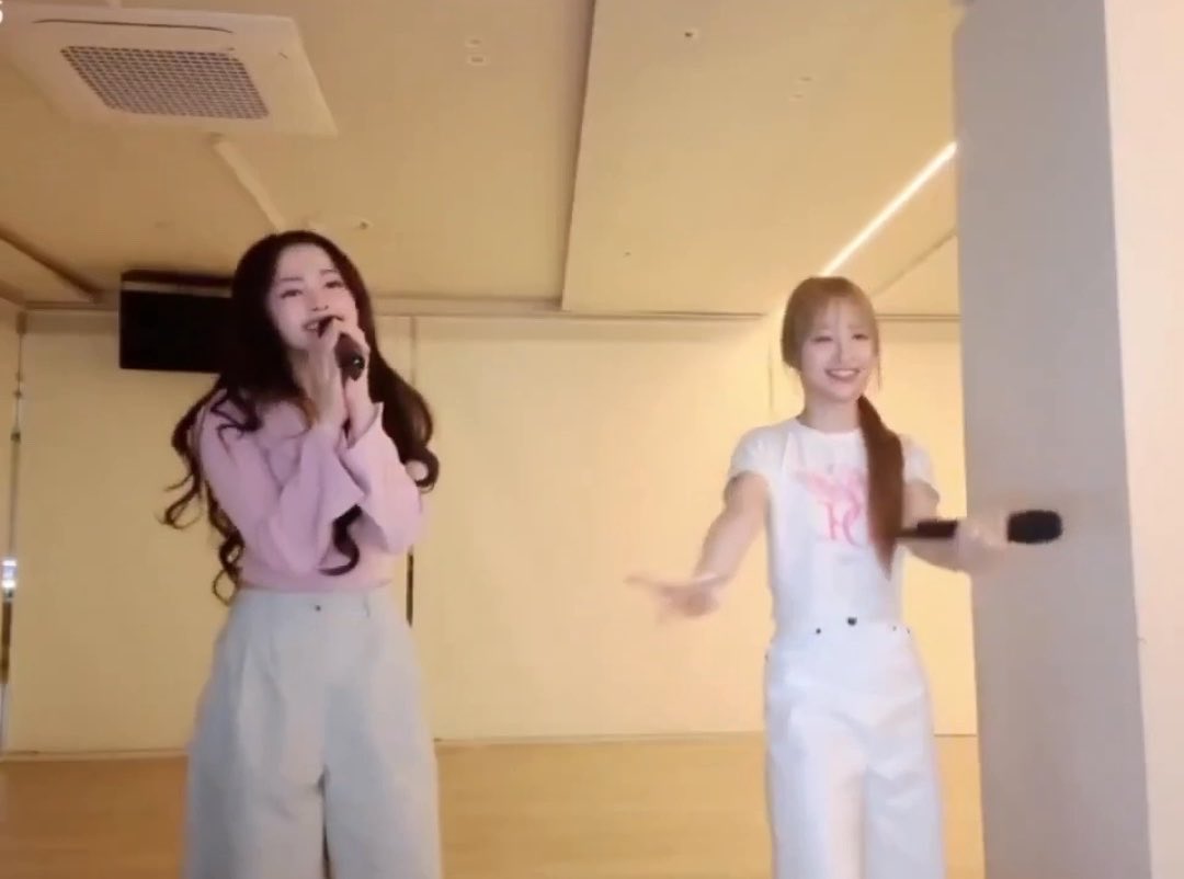Two young women in a dance studio, one in a pink shirt holding a microphone and the other in white holding a mic stand, appearing to rehearse.