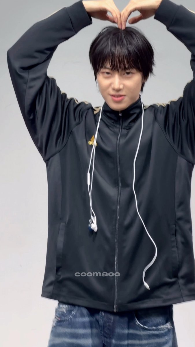 A young man with dark hair, wearing a black Adidas track jacket and white wired earphones, posing playfully with his hands forming a heart shape above his head; the image has a watermark 'coomaoo'.