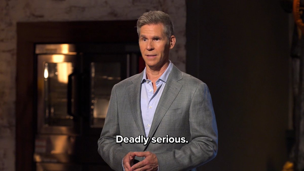 A gray-haired man in a gray suit and blue shirt speaks in a dimly lit room, with the subtitle "Deadly serious." displayed on screen.