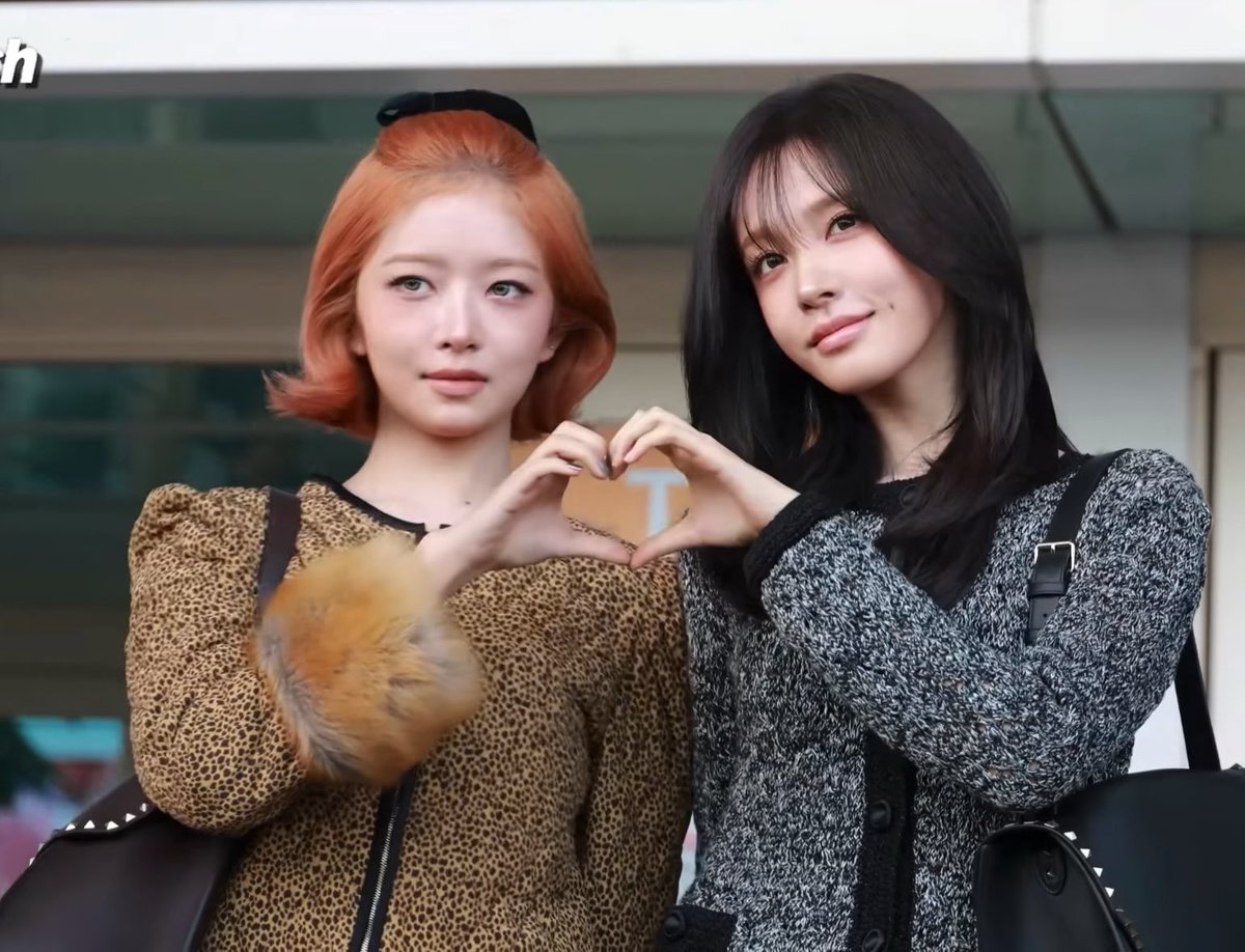 Two young women posing together outdoors, making a heart shape with their hands; the left has orange hair in a leopard print coat with a furry cuff, and the right has long black hair in a gray knit cardigan.