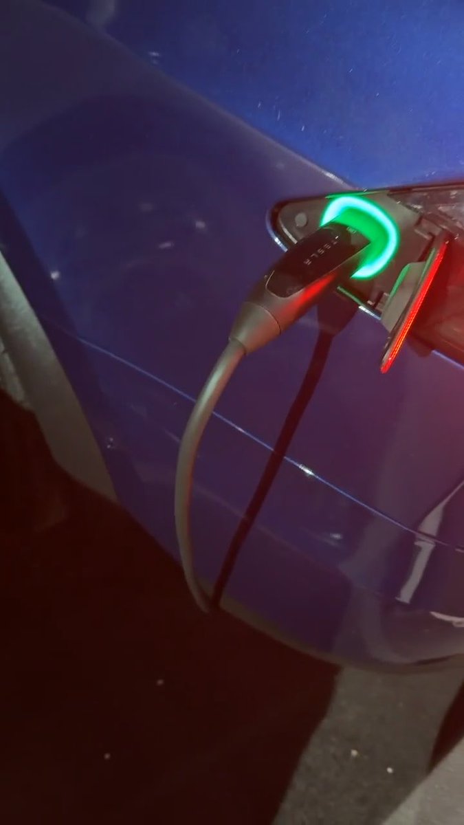 A Tesla EV charging cable plugged into a blue vehicle port, with the connector glowing green and red, and 'TESLA' branding visible on the plug.
