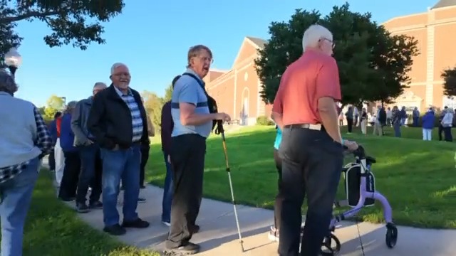A long line of attendees, including elderly individuals using walkers and canes, stretches along a sidewalk outside a brick building, waiting to enter a sold-out event featuring Heather Cox Richardson and a speaker, with over 700 Iowans expected to attend.