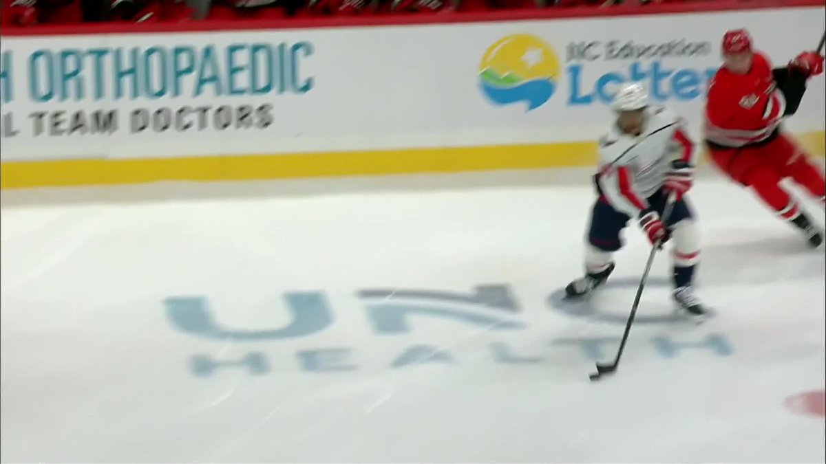 A hockey player in a white uniform skates with the puck on the ice rink while another player in red pursues, with 'USA HEALTH' visible on the ice and 'BC Orthopaedic Team Doctors' and 'BC Lottery' advertisements on the boards.