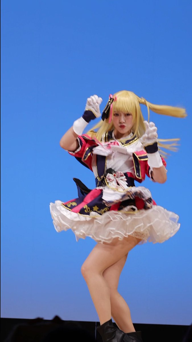 A cosplayer dressed as a blonde anime character in a colorful pink, white, and navy idol costume with a frilly skirt and star-patterned details jumps mid-performance against a bright blue backdrop. The post references "推しの子" (Oshi no Ko) and tags "雷電さん" and "ミクロンズ," suggesting this is a cosplay performance clip, with "コスパフォ" indicating a costume photoshoot or stage event.