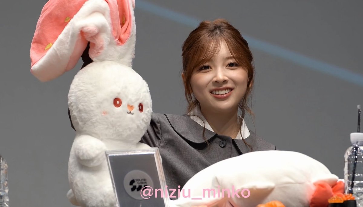 NiziU's MIIHI smiles brightly while holding up a white bunny plush with large pink ears at what appears to be a fan event, recalling the rabbit she gave during her Korean debut three years ago. The watermark "@nizu_minko" is visible in the bottom right corner of the image.