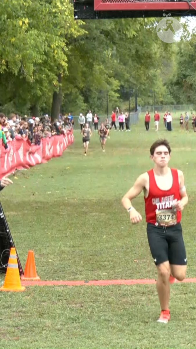 A young male runner wearing a red 'Carl Albert Titans' jersey with bib number 3374 crosses what appears to be a finish line at a cross-country race, with spectators behind red barriers and other runners visible in the background.