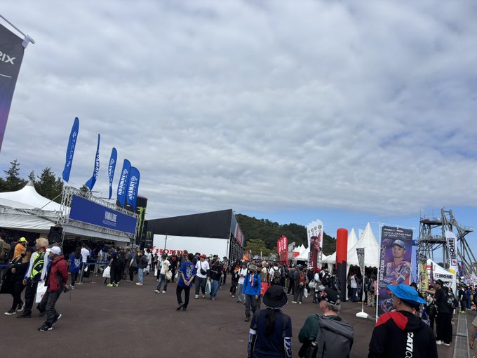 An outdoor event scene with crowds of people walking among white tents, blue flags, and banners including Honda and FOX branding, with cloudy skies and mountains in the background.