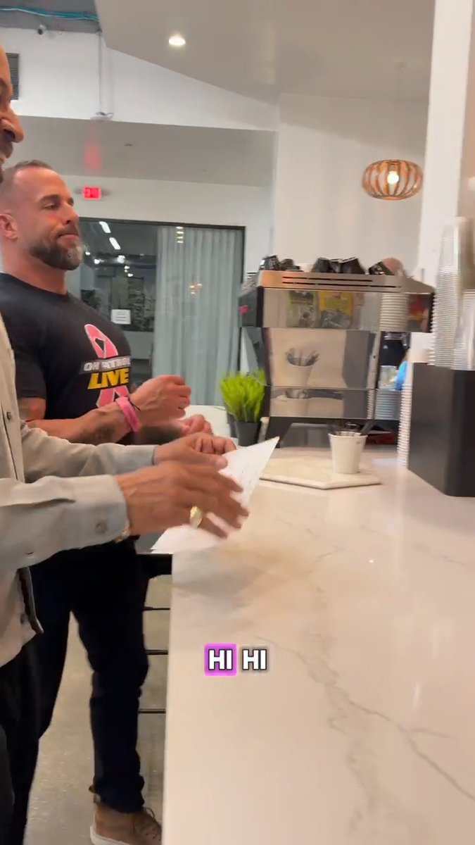 Two men stand at a bright marble coffee counter, one in a black "ON PATROL LIVE" t-shirt, exchanging a friendly greeting of "HI HI" (displayed as text overlay) in a casual café setting. The image ties to the post by showing the "On Patrol: Live" branding on the shirt, connecting to the show being promoted.