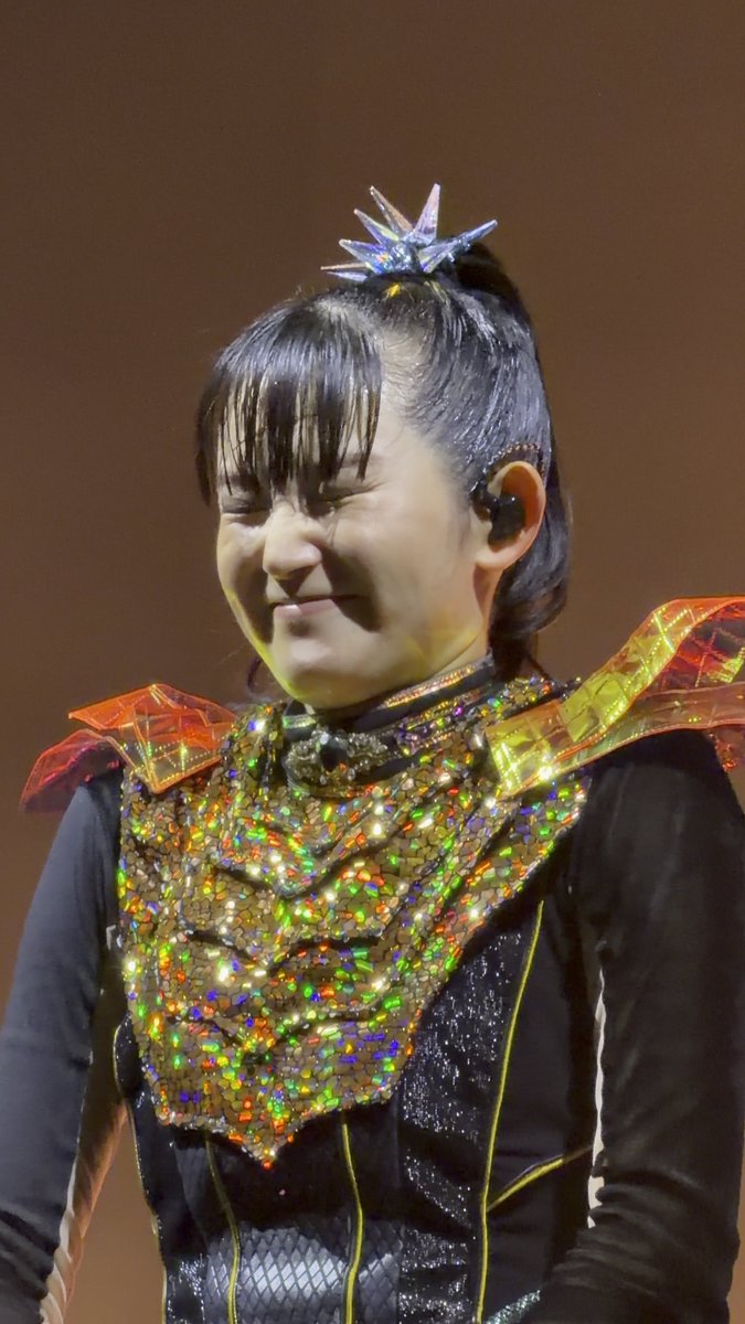 A performer on stage in an elaborate costume featuring a holographic, sequined chest plate, orange translucent shoulder accents, and a crystal-like star hair ornament, captured with an expressive face.