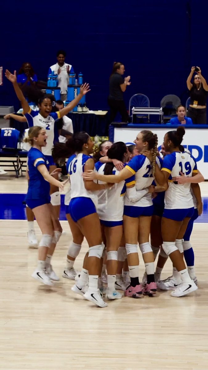 A women's volleyball team in blue and white uniforms, including player #16, #21, and #19, celebrate together in a huddle on a court with a coach clapping in the background.