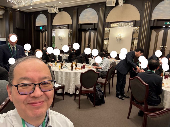 A selfie taken at a formal banquet dinner event with multiple attendees seated at round tables in suits; faces of other people are obscured by white circle stickers.