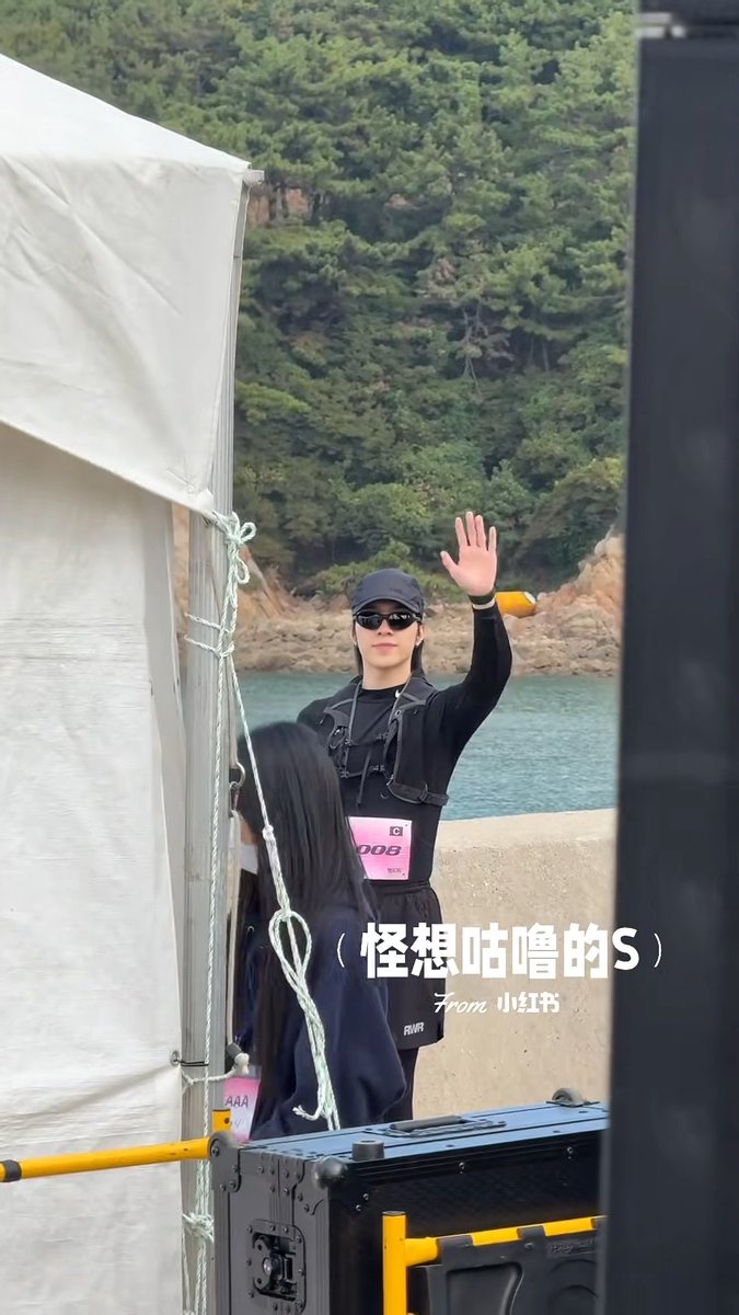 A man wearing a black cap, sunglasses, and a "556" race bib waves his hand near a white tent by the waterfront, with another person wearing bib "444" standing nearby; Chinese text on the image reads "怪想咕噜的S" with a "From 小红书" watermark, matching the post about Hendery at the SMRUN event.