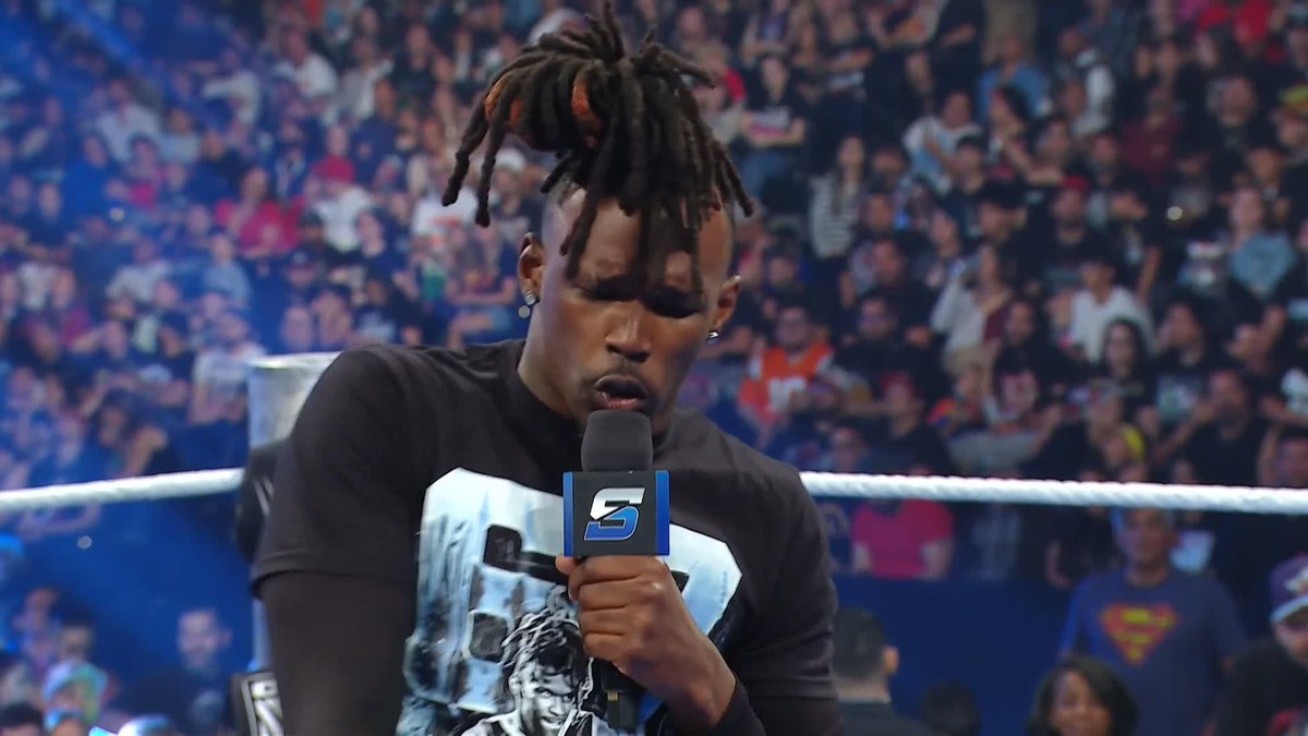 A wrestler with dreadlocks, wearing a black graphic tee, speaks into an "SmackDown" mic inside a wrestling ring surrounded by a packed arena crowd, with a Superman shirt visible among the spectators. The caption from the post appears to reference a promo segment critiquing a rival's lengthy journey to a WWE title, with the wrestler likely delivering the cutting line.