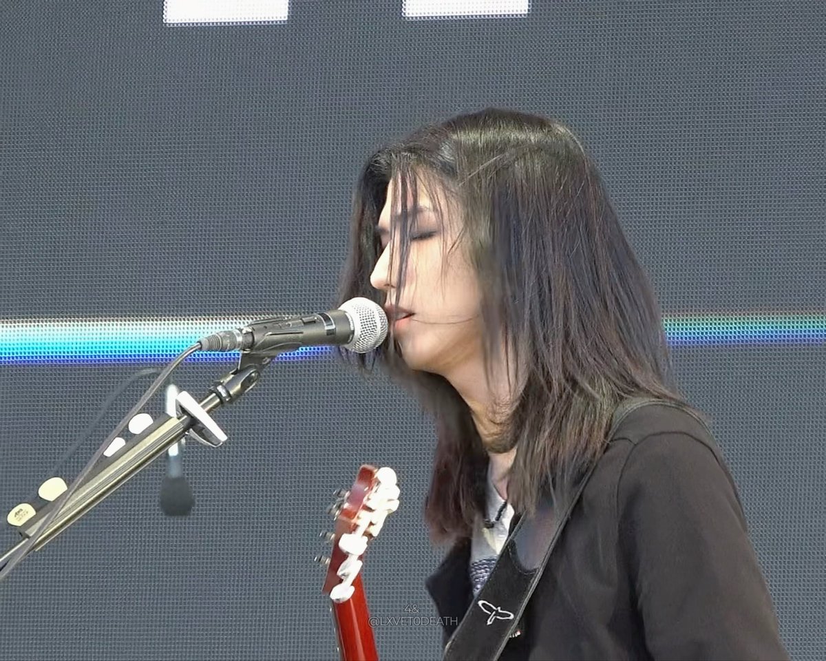 A side profile of a female musician with long dark hair singing into a microphone while playing an electric guitar at an outdoor concert, with a watermark reading '@LXVETODEATH'.