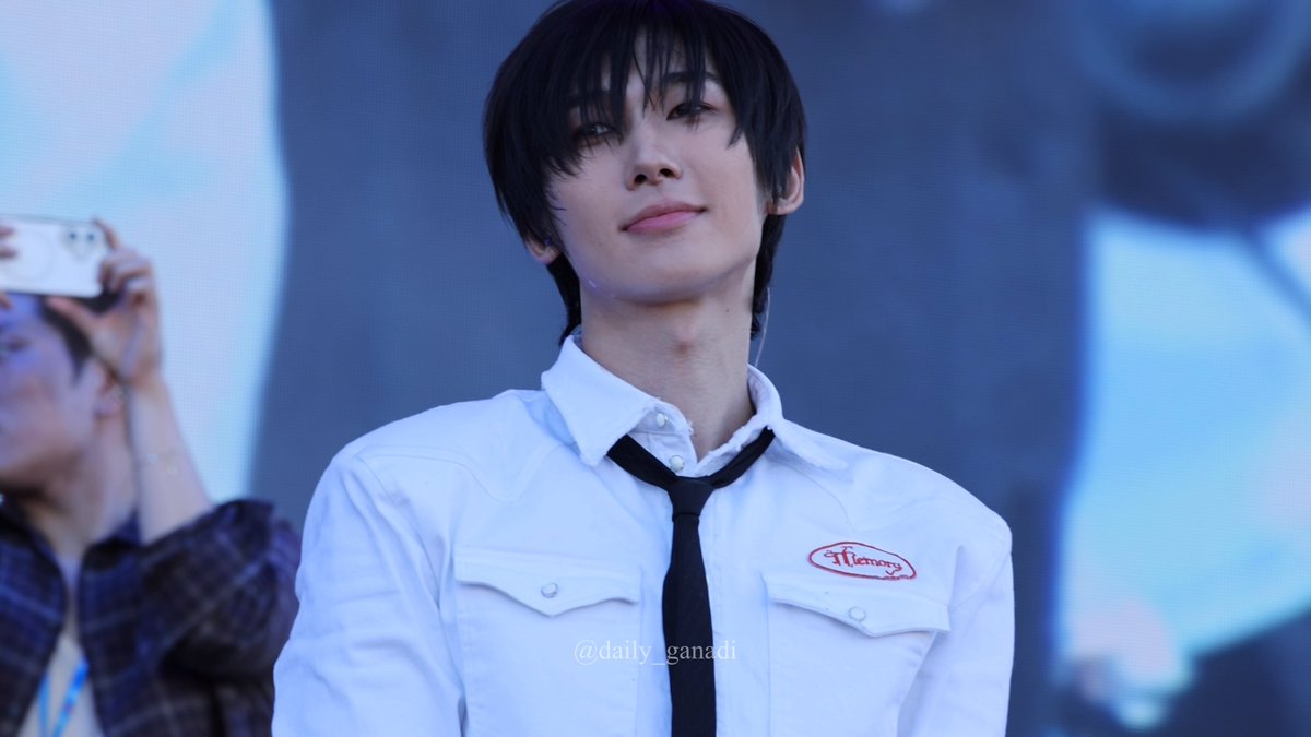 A young man with dark hair wears a white button-up shirt with a black tie and a 'Memory' patch, smiling softly at the camera with the watermark '@daily_ganadi'.