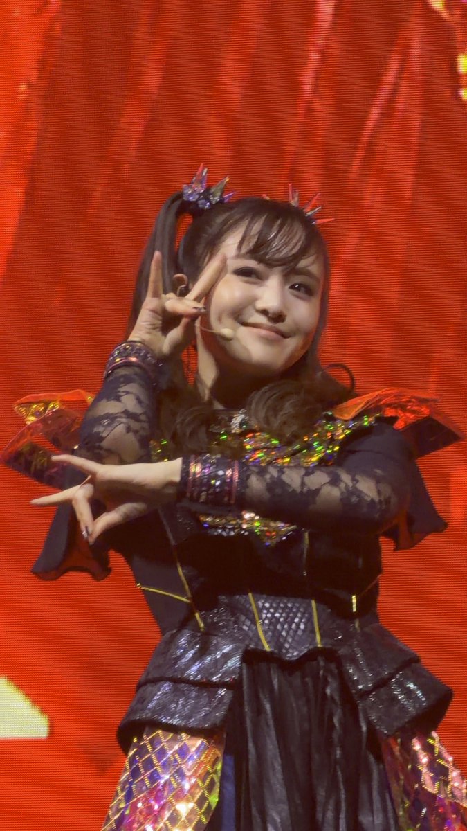 A young woman in an elaborate black gothic-style outfit with colorful sequins and a crown-like headpiece smiles while making peace signs on stage against a red backdrop.