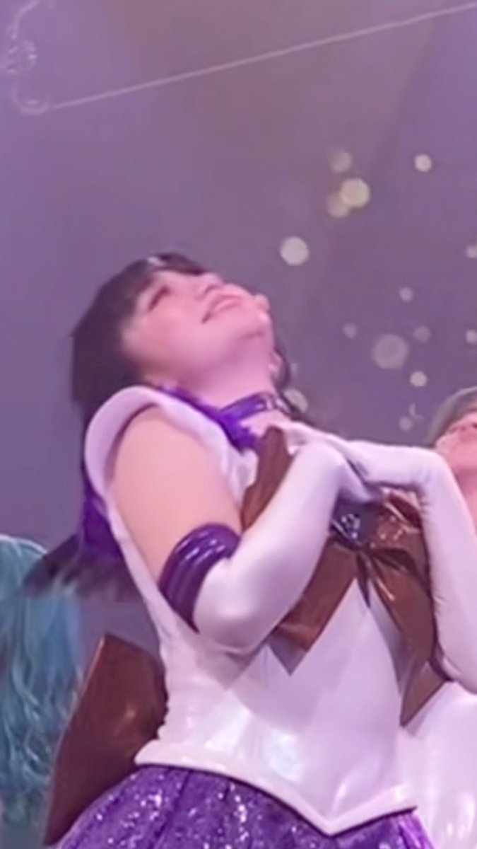 A performer dressed as Sailor Saturn (Tomoe Hotaru) from Sailor Moon in a white and purple costume, leaning back with eyes closed on what appears to be a stage.
