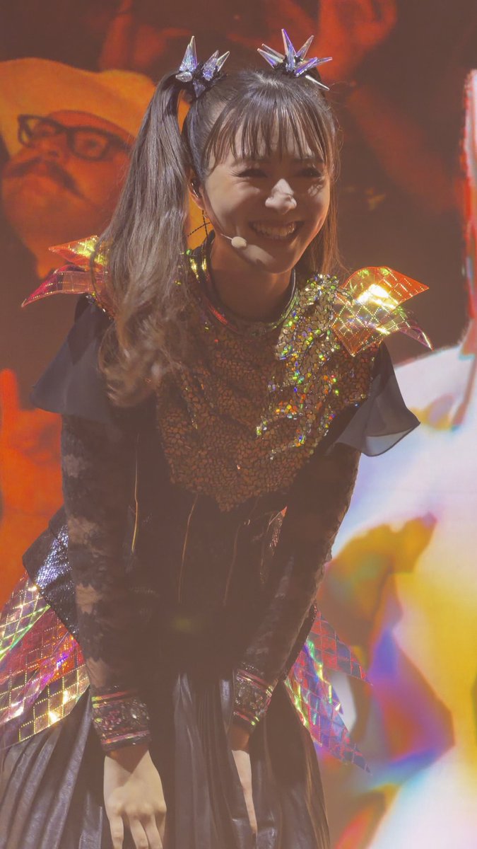 A smiling performer wearing a black dress with iridescent spiked shoulder accents and star hair accessories poses on stage under warm lighting.