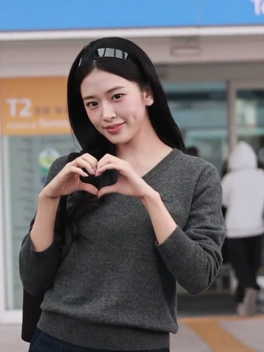 A young woman with long black hair and a gray sweater makes a heart shape with her hands while smiling at the camera, likely at an airport (T2 visible).