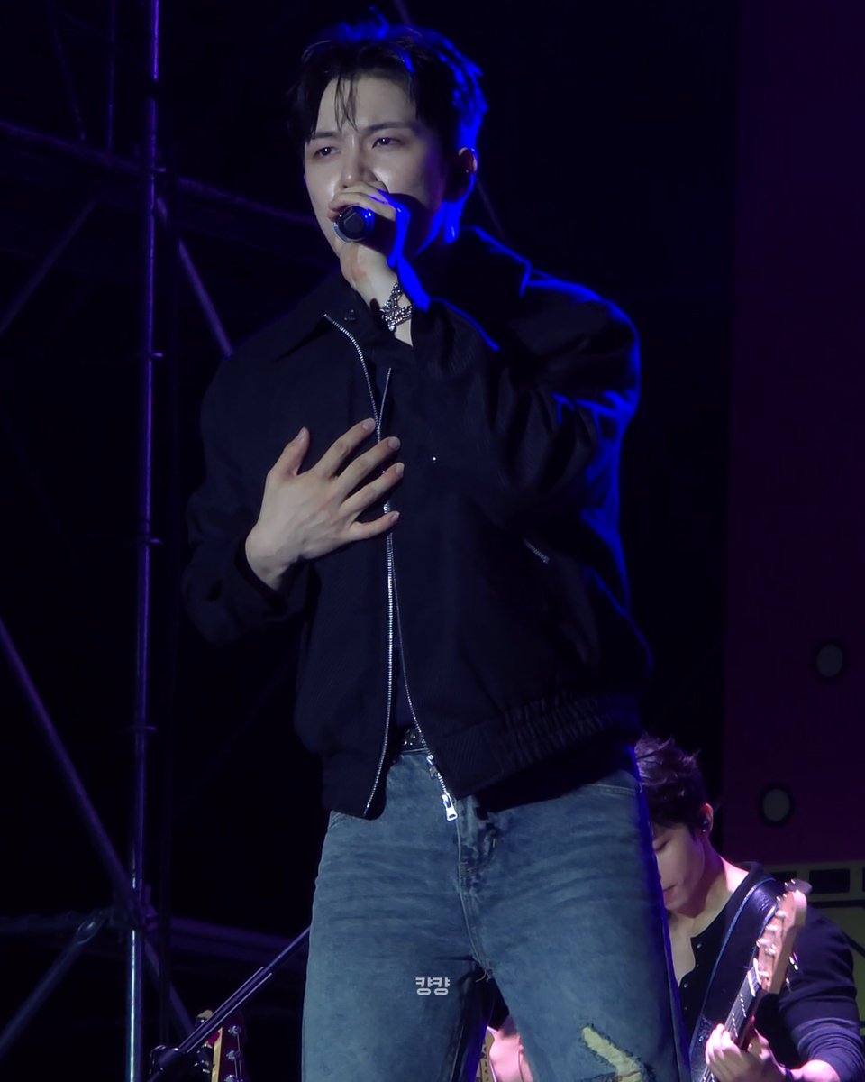 A performer on stage in a black jacket and jeans singing into a microphone, with blue stage lighting and another musician with a guitar in the background.