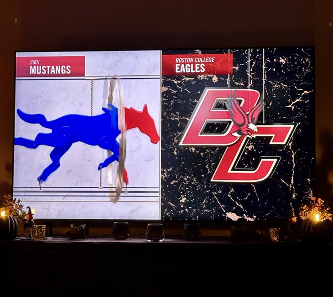 A TV screen displays a college sports matchup graphic between 'SMU MUSTANGS' featuring a blue horse logo and 'BOSTON COLLEGE EAGLES' with their BC eagle logo, with small pumpkins and candles arranged below.