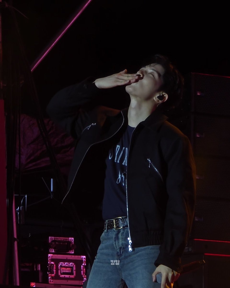 A young performer on stage in a dark setting with pink lighting, wearing a black bomber jacket over a graphic t-shirt and jeans, looking upward while holding a microphone.