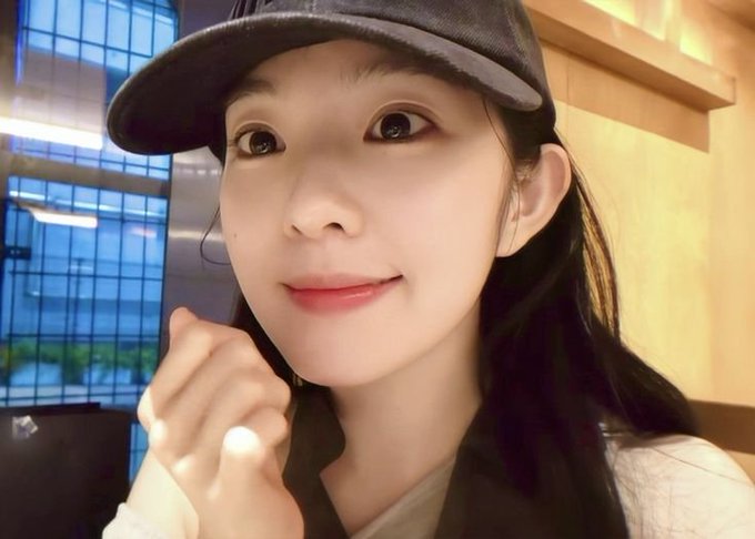 A close-up selfie of a young woman wearing a dark gray baseball cap and a beige sweater, looking at the camera with a slight smile in what appears to be an indoor setting.