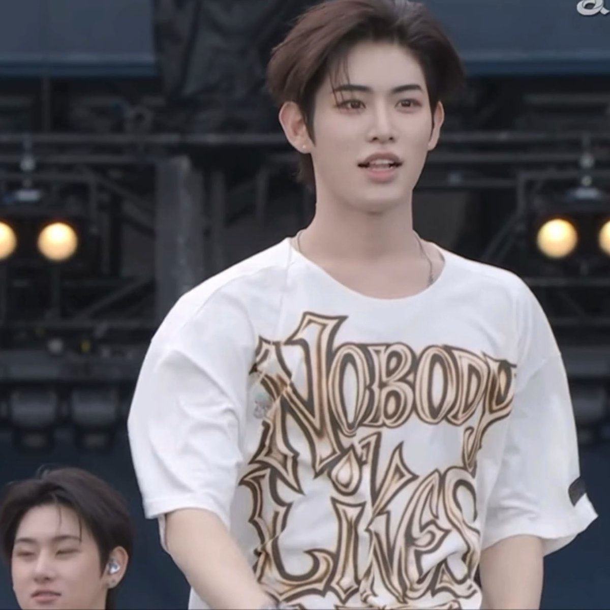 A young man with brown hair wearing a white graphic t-shirt with 'NOBODY' printed on it, appearing to be at an outdoor event.
