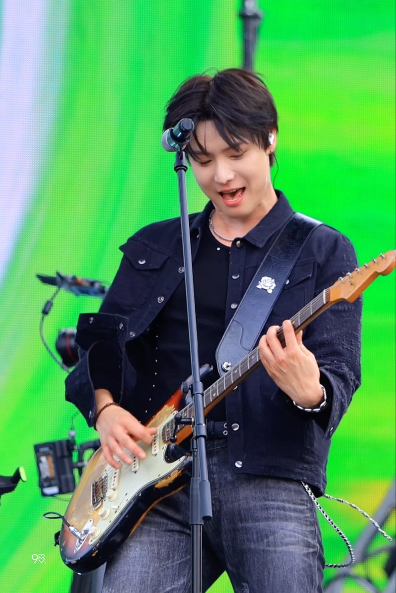 A male musician with dark hair playing an electric guitar and singing into a microphone on stage with a bright green background.