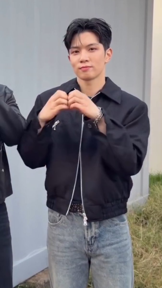 A photo of a young man with dark hair wearing a black zip-up jacket and light blue jeans, making a heart shape with his hands and smiling at the camera outdoors.