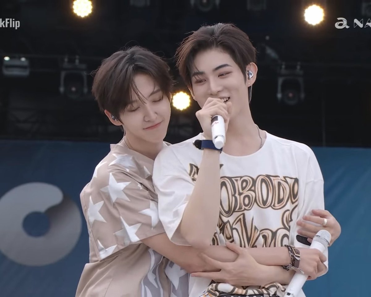 Two young male performers are on stage, with one embracing the other from behind while singing into a microphone; both are smiling, with one wearing a beige star-patterned shirt and the other a white graphic t-shirt.