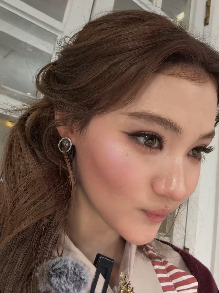 A close-up selfie of a woman with brown hair, wearing winged eyeliner, pink blush, and a silver oval earring.