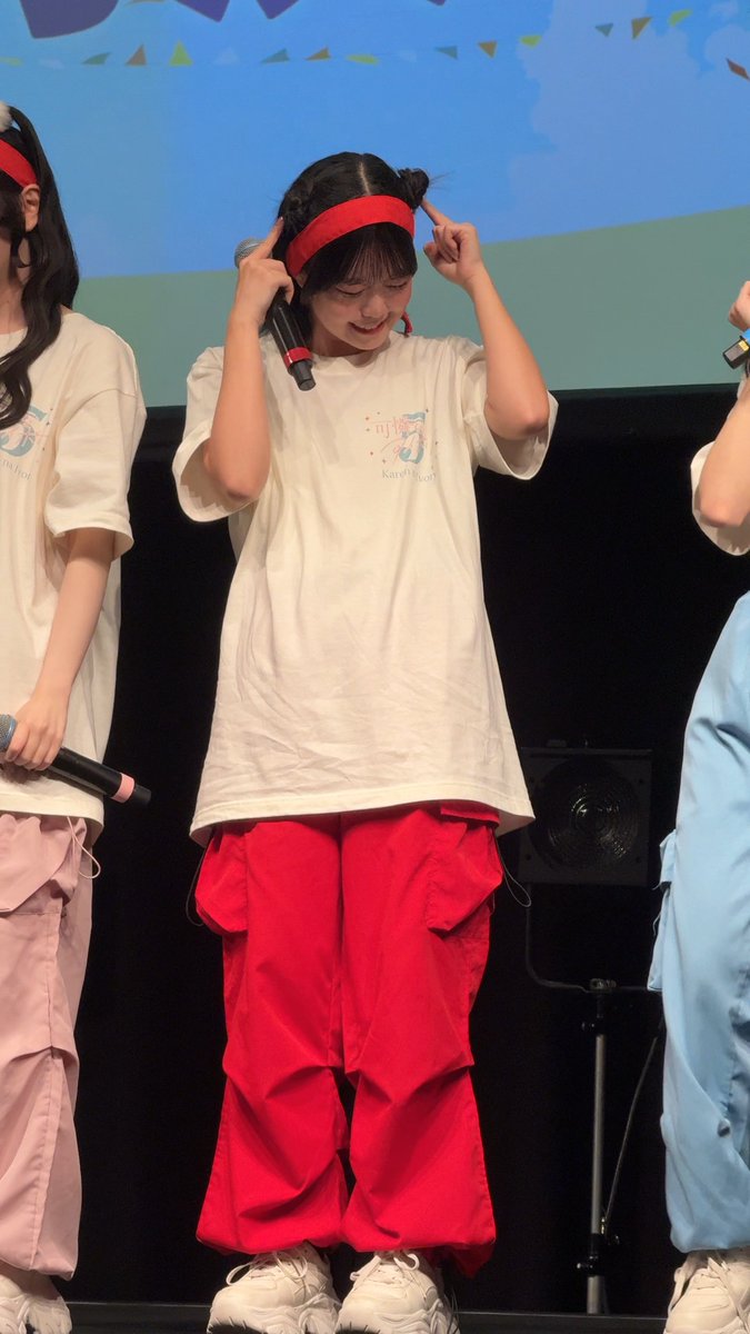 A performer on stage wearing a red headband, white t-shirt, and baggy red cargo pants holds a microphone while making a playful gesture with her fingers near her temples.